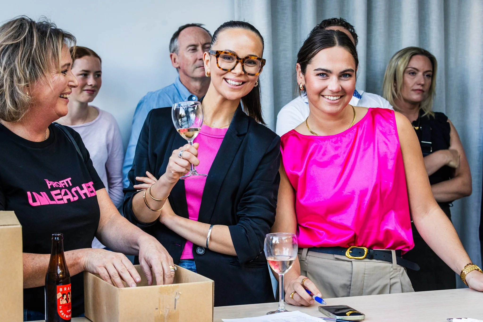Group of people at a social event, some holding wine glasses, smiling and engaging with each other, with a table and gray curtains in the background.