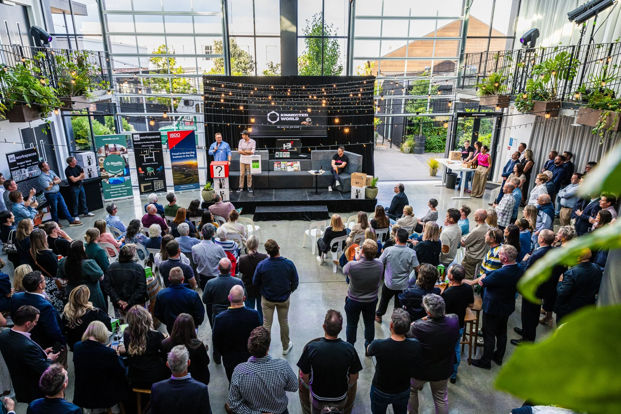 A large indoor event with a stage and many attendees watching a presentation. The stage has a black backdrop with string lights and various banners, and several speakers are on stage, some standing and some seated. The audience is seated and standing