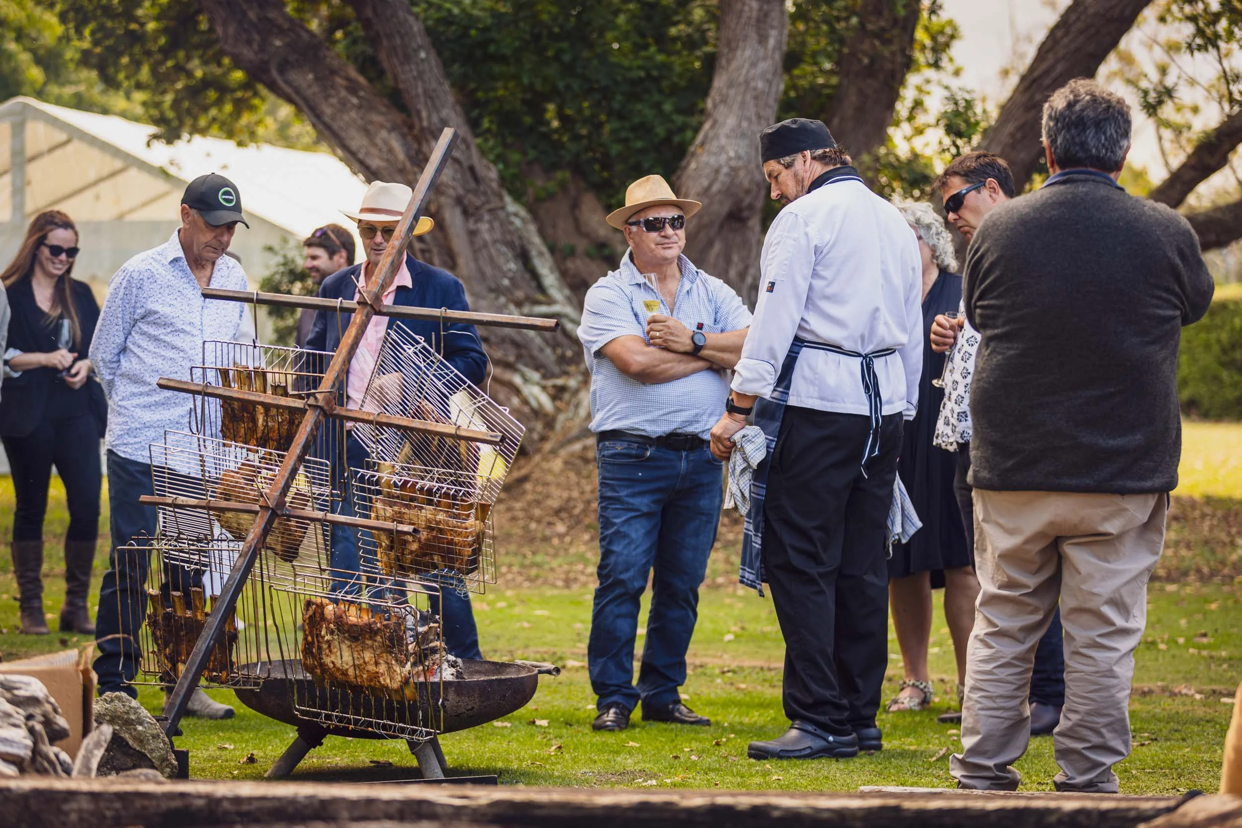 People gathered outdoors around a barbecue grill with meats cooking, under large trees with a white tent in the background.