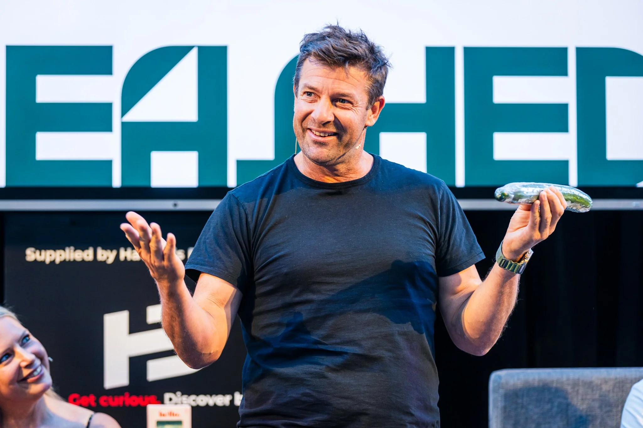 Man speaking at a presentation, holding an object that looks like a cucumber, with large text reading 'HACKED' in the background.