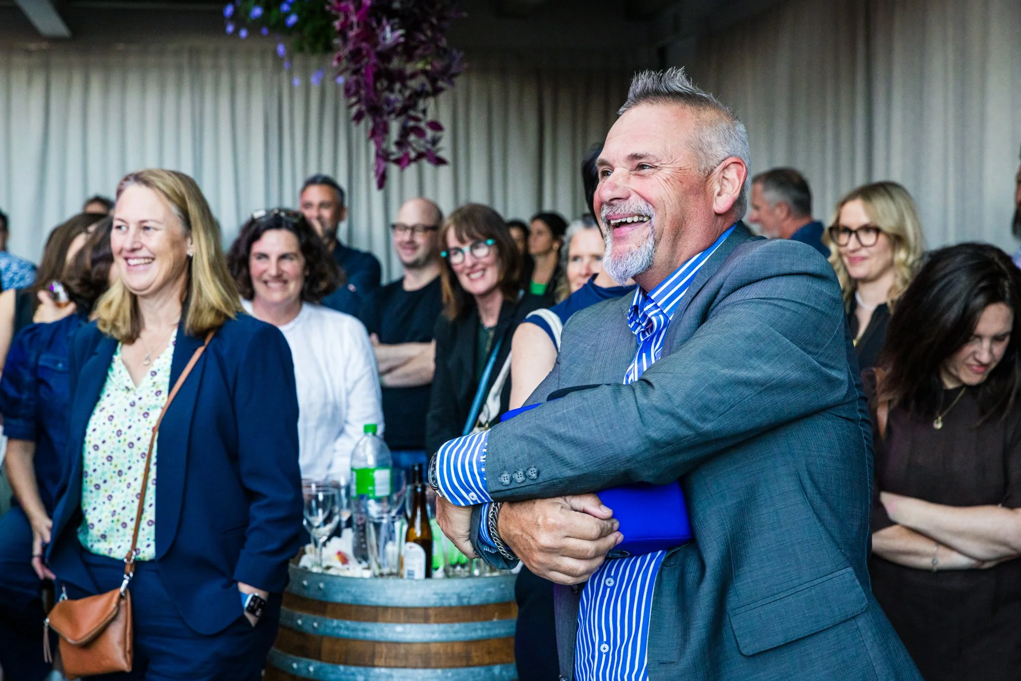 A group of people at a social event, smiling and laughing, with a man in a gray suit and striped blue shirt in the foreground.