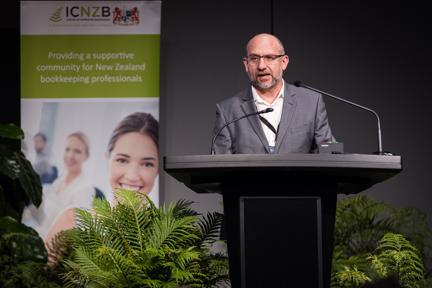 A man in a gray suit speaking at a podium during a conference, with a banner behind him that reads "Providing a supportive community for New Zealand bookkeeping professionals" and shows images of smiling women, surrounded by green plants.