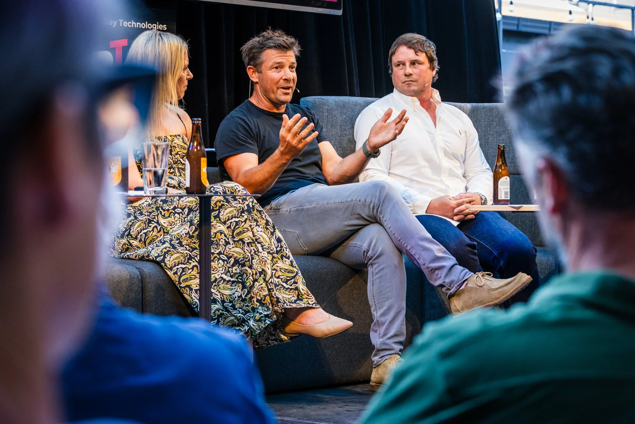 A panel discussion with three people speaking on stage, with an audience in front. The speaker in the middle is a man wearing a black t-shirt and gray pants, gesturing with hands. To his right, another man in a white shirt listens attentively. A woma