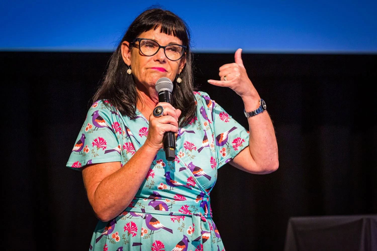 A woman with dark hair, glasses, and hoop earrings speaking into a microphone on stage, making a hand gesture resembling a phone call, wearing a colorful dress with bird and floral patterns.