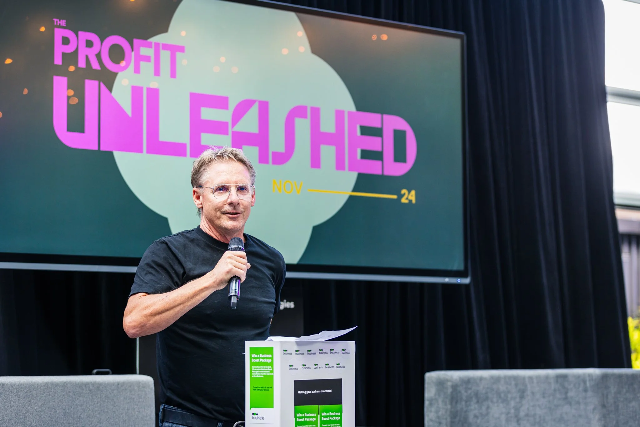 A man holding a microphone giving a presentation at an event titled 'Profit Unleashed' with a large screen behind him displaying the event name and date, November 24.
