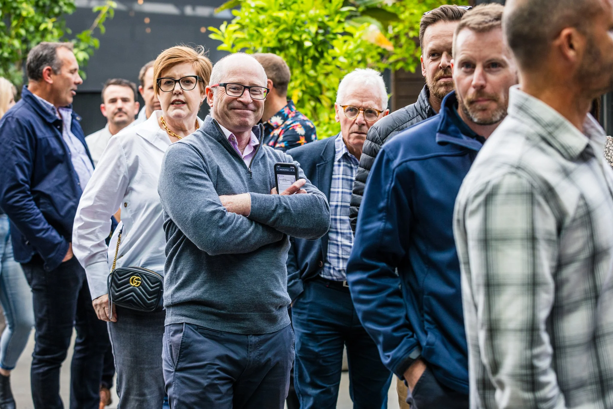 A group of people standing in a line outdoors, waiting or observing, with greenery in the background.