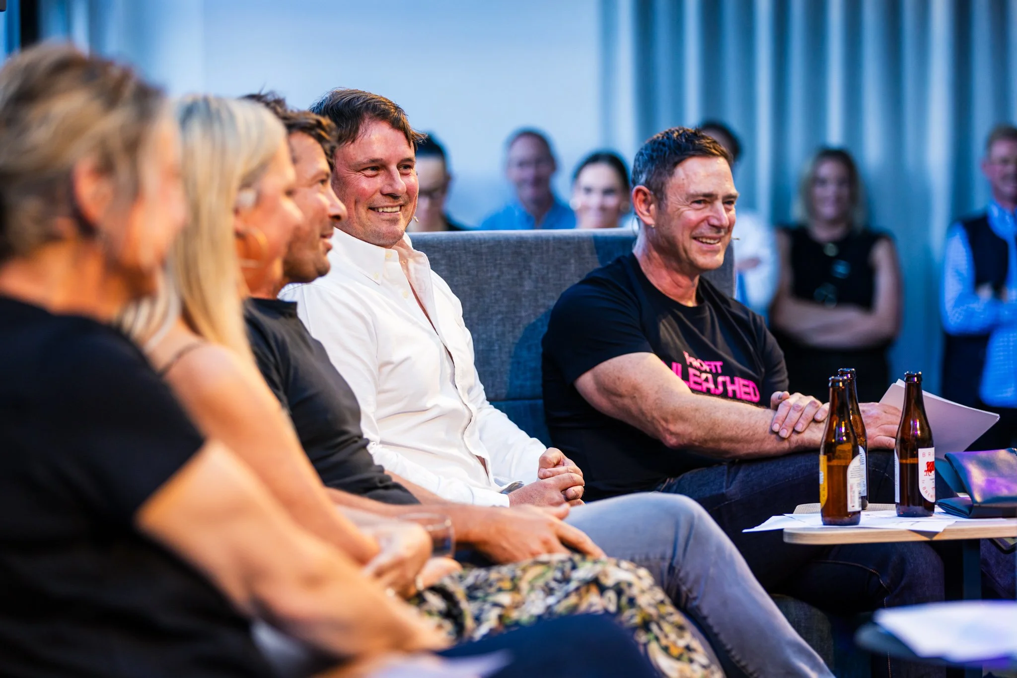 A group of people sitting in a row at an indoor event, smiling and engaging, with some standing in the background. Three men are prominently visible, with one wearing a black T-shirt that says 'Your Heart Led.'