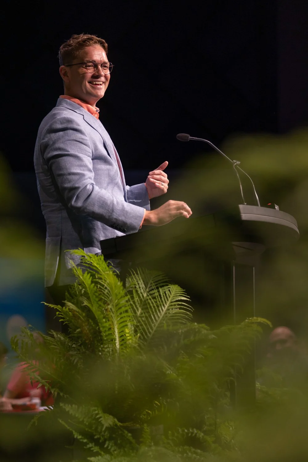 Man in gray blazer and glasses smiling while standing at a podium with microphone, speaking at an event, with green plants in the foreground.