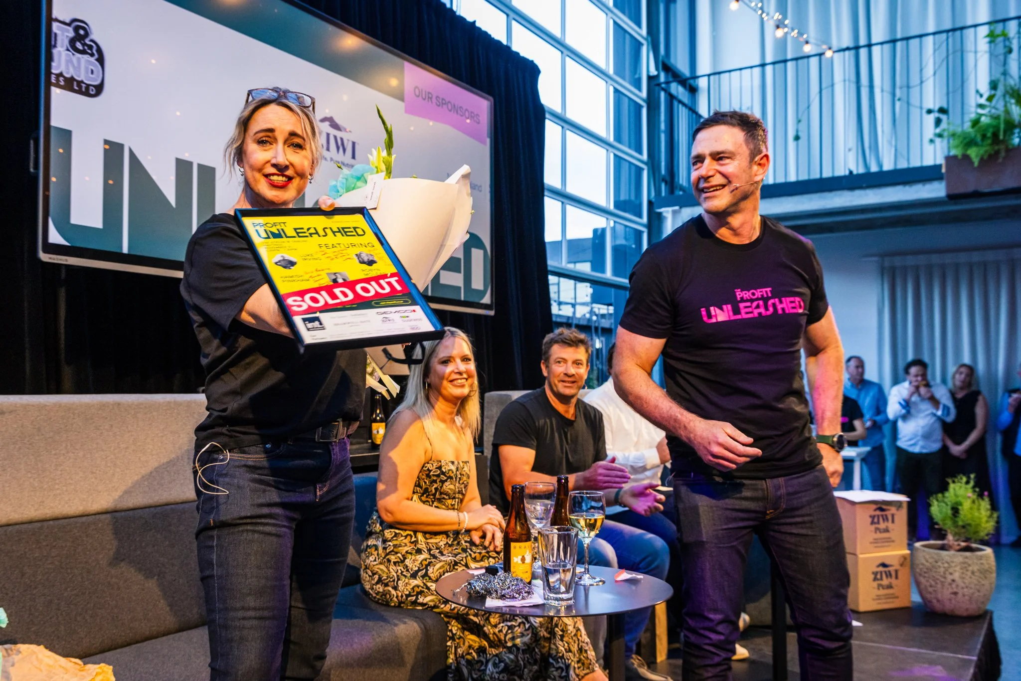 A woman holding a framed certificate and bouquet standing on stage at an event, with three seated people and a crowd in the background. The woman is smiling, and the certificate has a large red "SOLD OUT" label. The stage backdrop has logos and text,