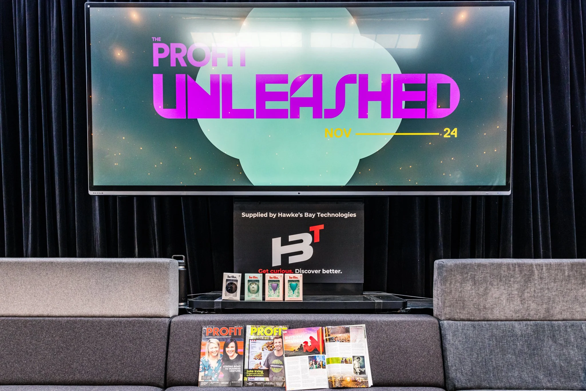 A conference or event room with a large screen displaying an advertisement for 'Profit Unleashed' happening on November 24. Below the screen, there is a smaller monitor with the Hawke's Bay Technologies logo and slogan, 'Get curious. Discover better.