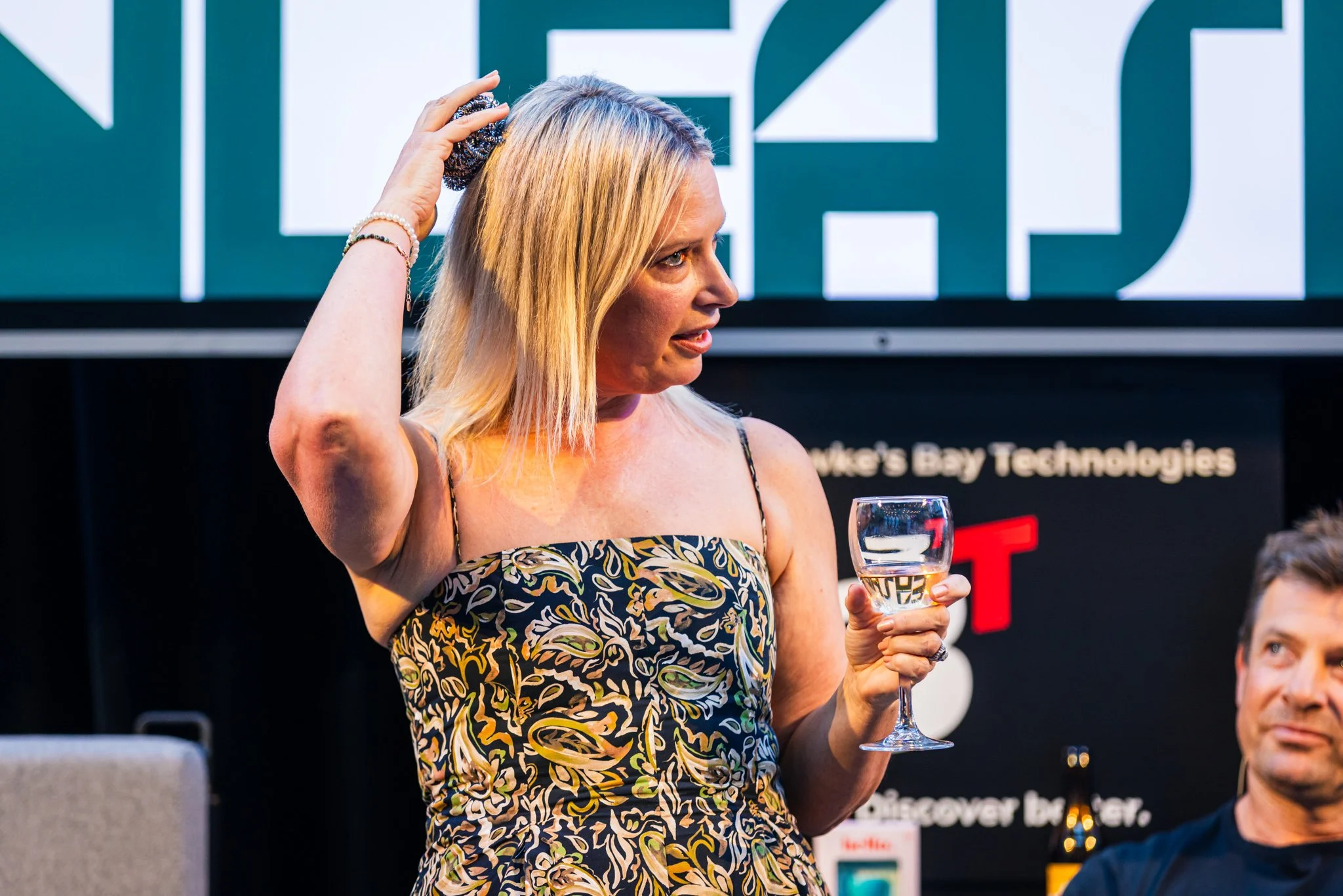 Blonde woman with a sleeveless patterned dress holding a glass of water and touching her hair, sitting at a table during a discussion or presentation.