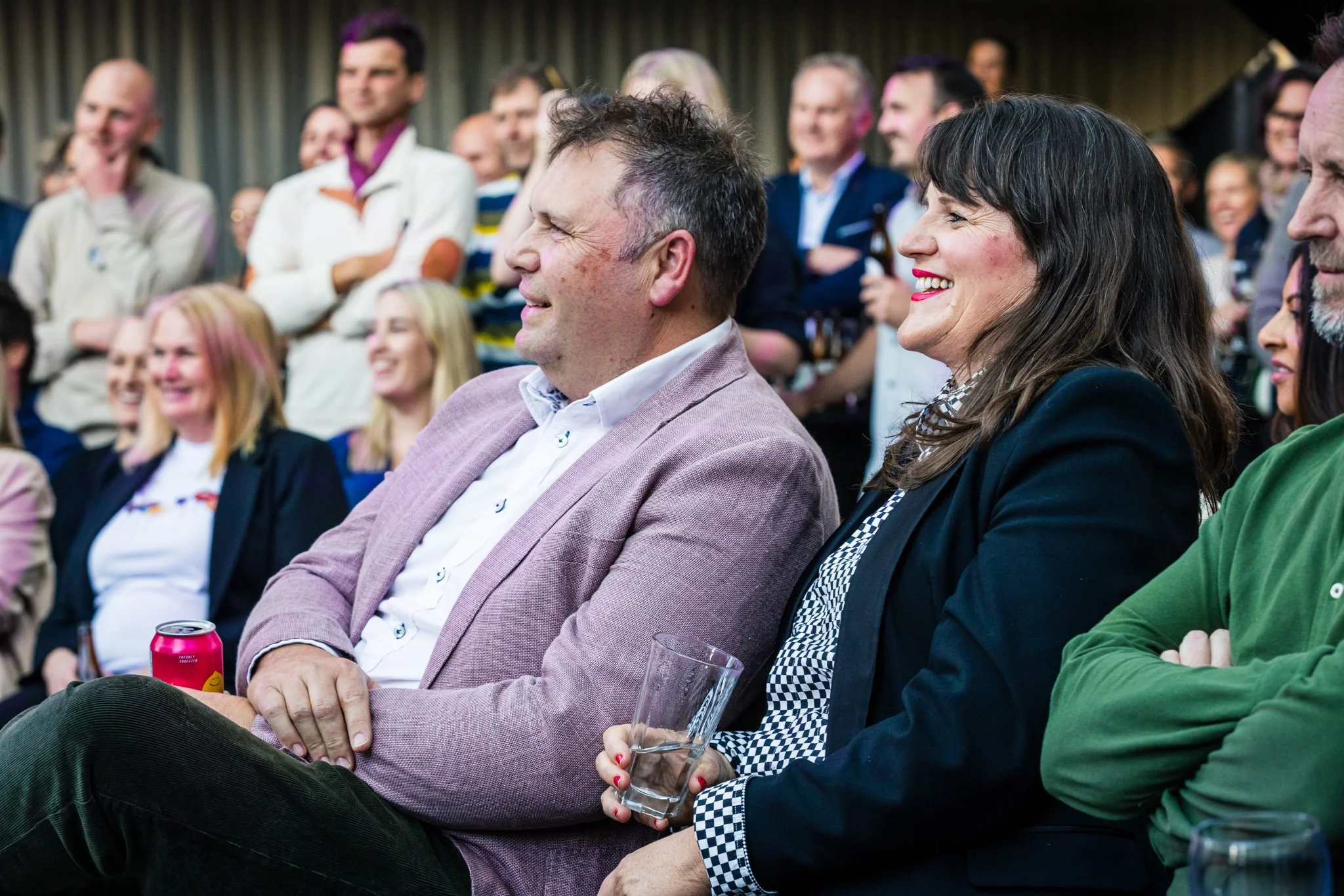 People attending a live event, sitting in chairs, smiling and laughing, some holding drinks, in an indoor setting with a curtain background.