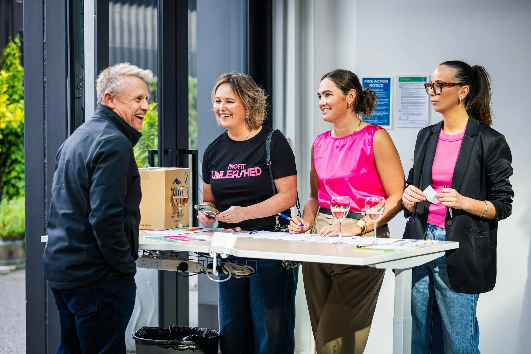 Four women standing at a registration table, talking and smiling at a man. The table has glasses of wine, papers, and a box on it.