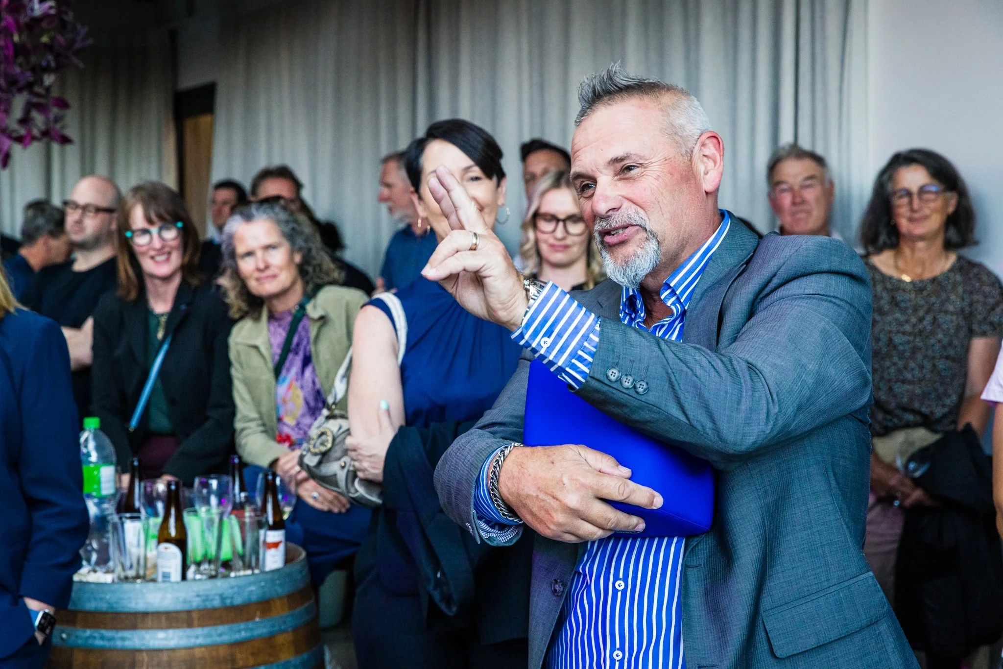 A man in a gray suit with a blue striped shirt holding a folder and gesturing while surrounded by a diverse group of people at a social gathering or conference.