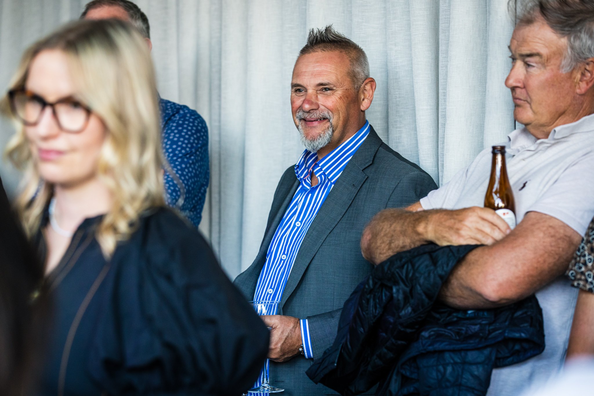 Group of people attending a social gathering indoors. A man in a gray suit with a blue striped shirt is smiling, and another man with gray hair is holding a beer bottle and crossing his arms.