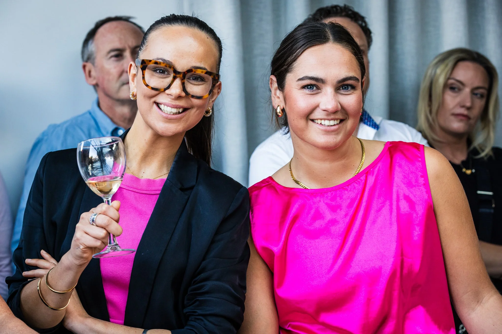 Two women smiling at a social gathering, one holding a glass of white wine, the other in a pink top, with other people in the background.