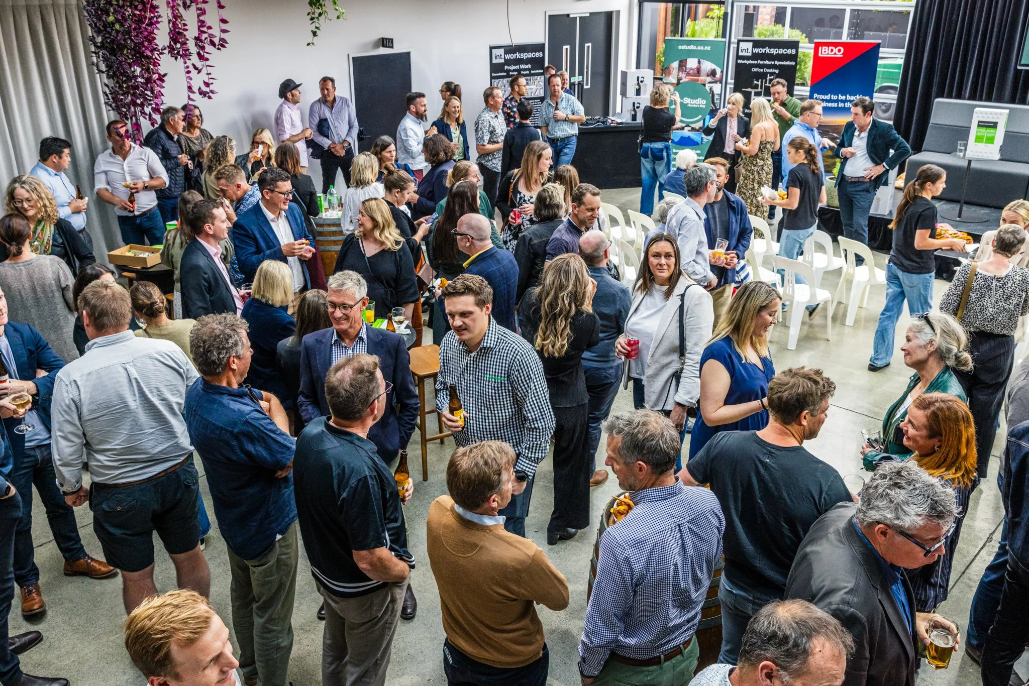 A large indoor networking event with many people talking, holding drinks and food, in a spacious room with banners, a stage, and seating areas.