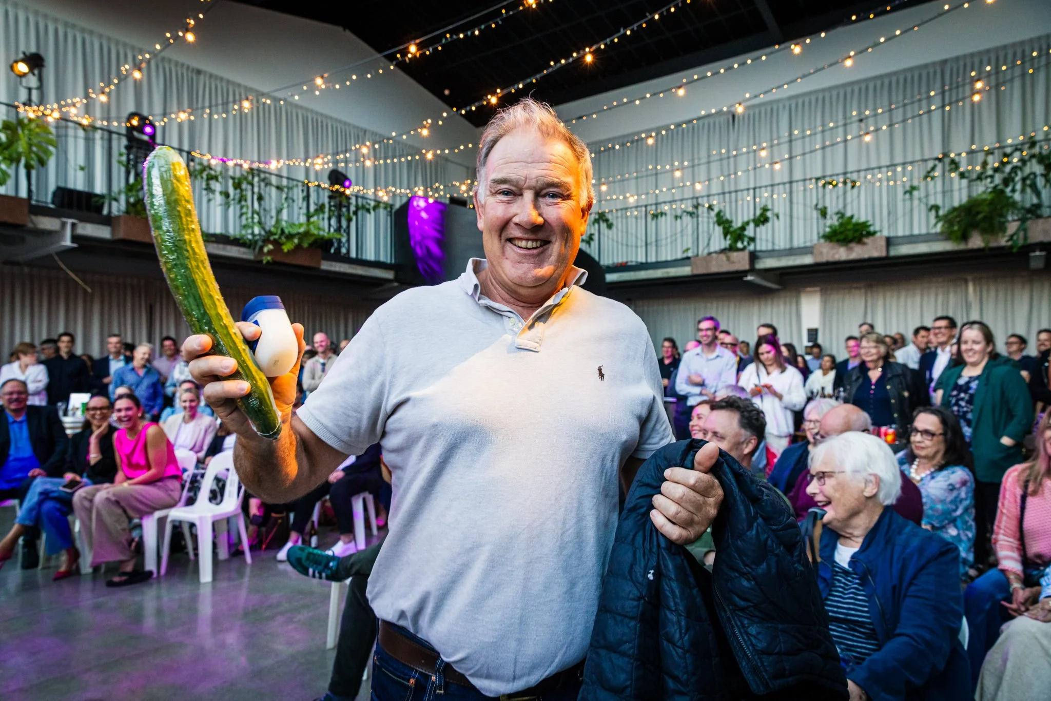 A man smiling at the camera while holding a pickle with a small container in it at an indoor event with multiple people seated and standing, decorated with string lights and plants.