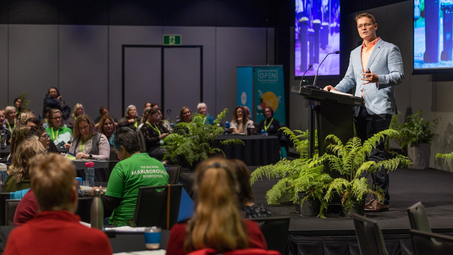 A man in a light gray blazer and orange shirt is speaking at a podium during a conference. The audience consists of women and men seated at round tables, some taking notes. There are large screens behind him displaying images, and potted ferns decor 