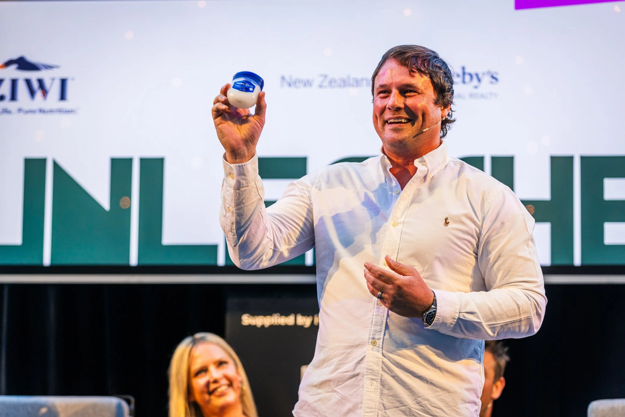 A man in a white shirt holding a small container of cream or lotion, standing on a stage with a large screen behind him displaying text and logos, smiling.
