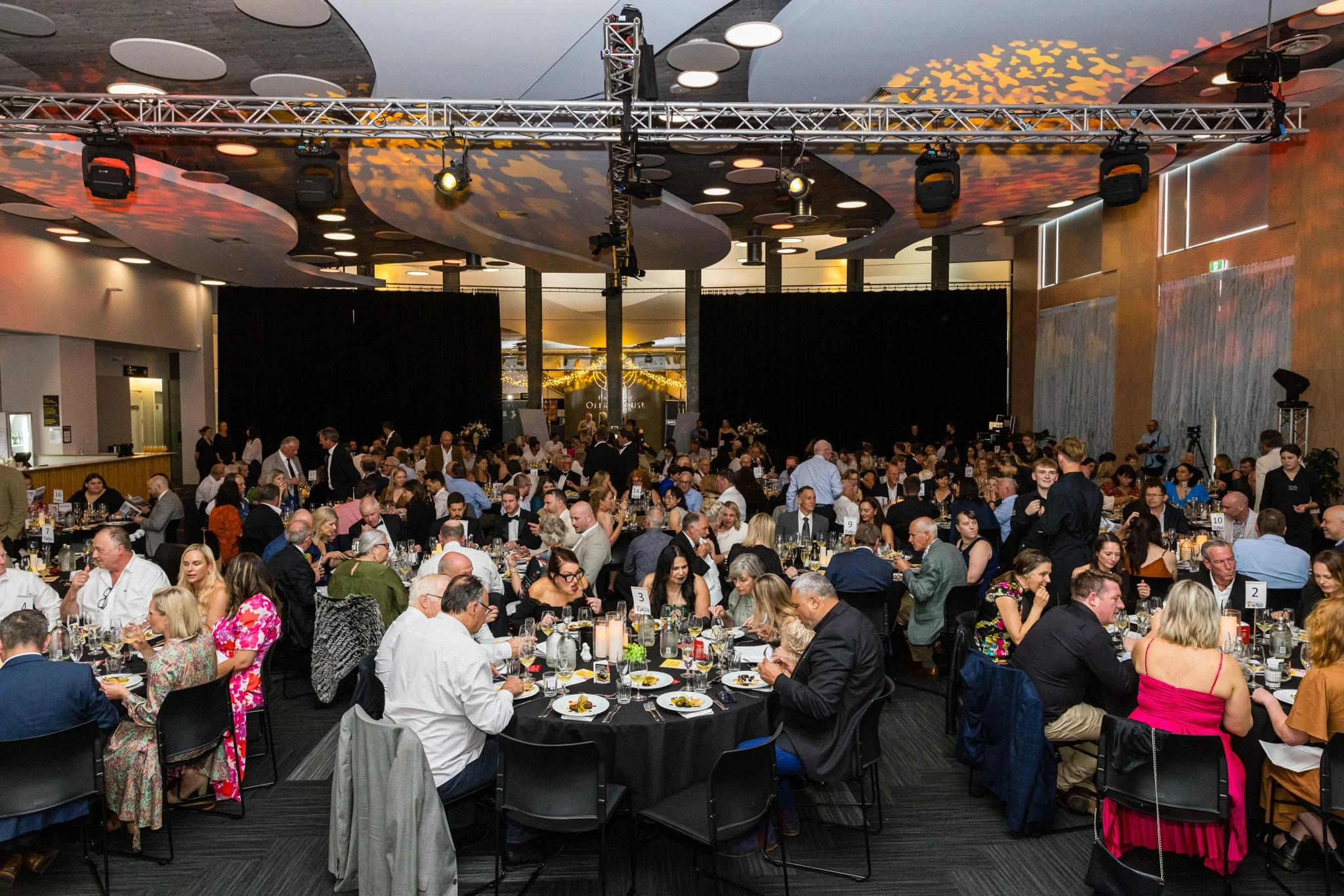A large banquet hall filled with round tables of people enjoying a formal event, with a stage at the front, black curtains, and decorative ceiling lighting.