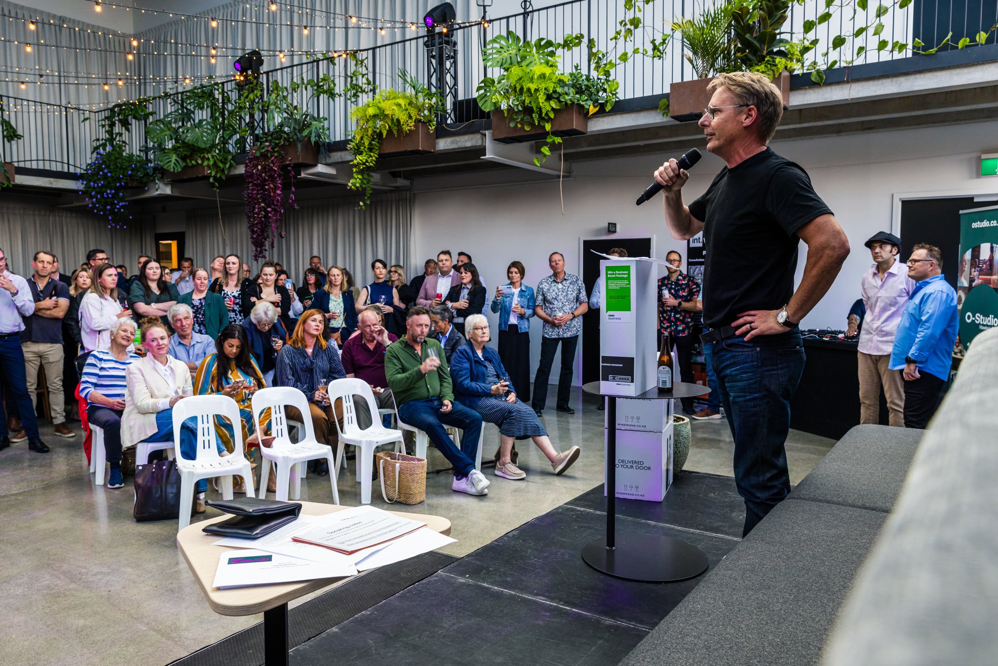 A man in a black t-shirt and jeans speaking into a microphone on a stage at a crowded indoor event. Audience members are seated and standing, listening attentively, with some taking pictures. The room has hanging plants and fairy lights.