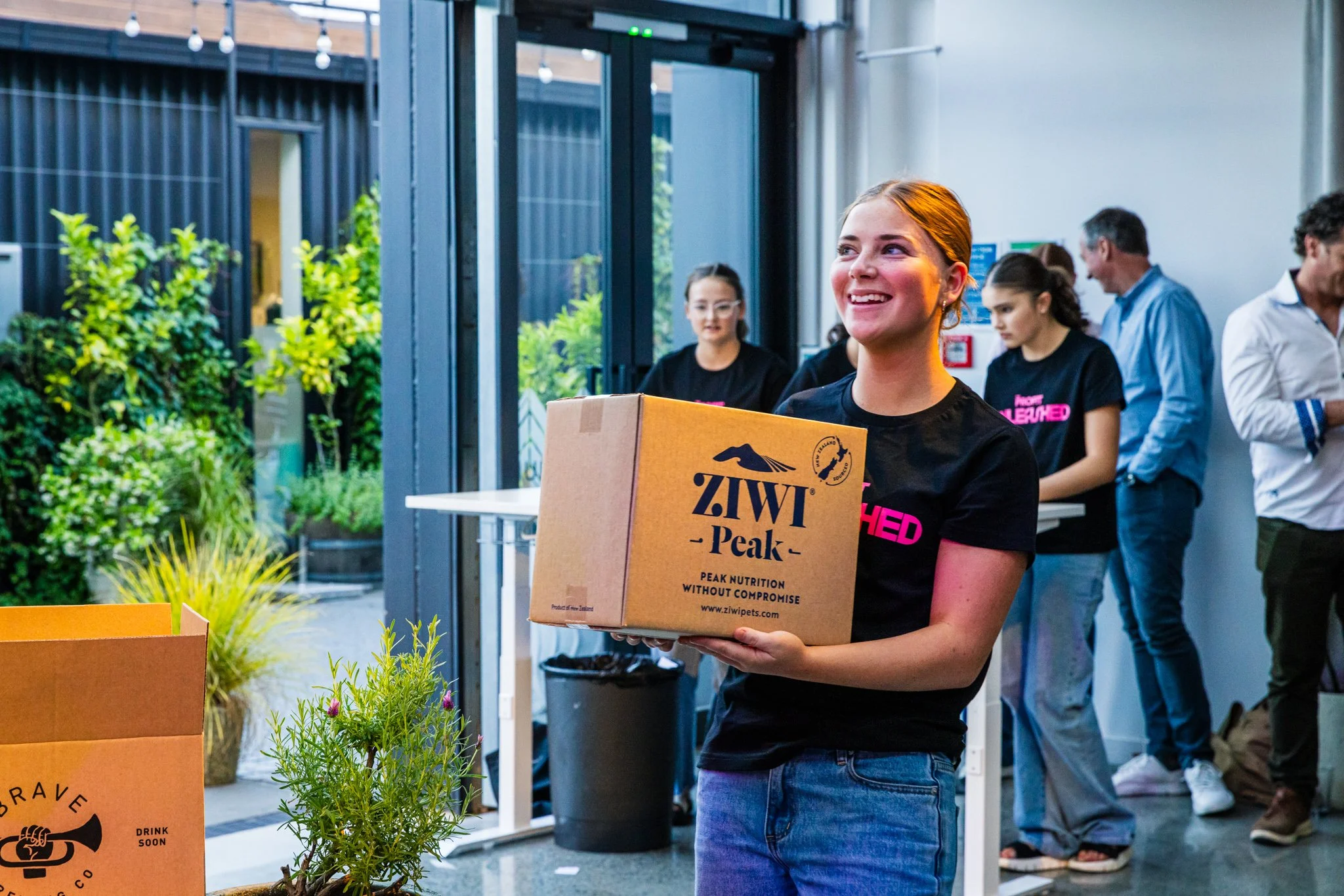 A young woman smiling and holding a box labeled ZIWI Peak at an indoor event, with others in the background including a woman in pink-printed black T-shirt and some people engaged in conversations.