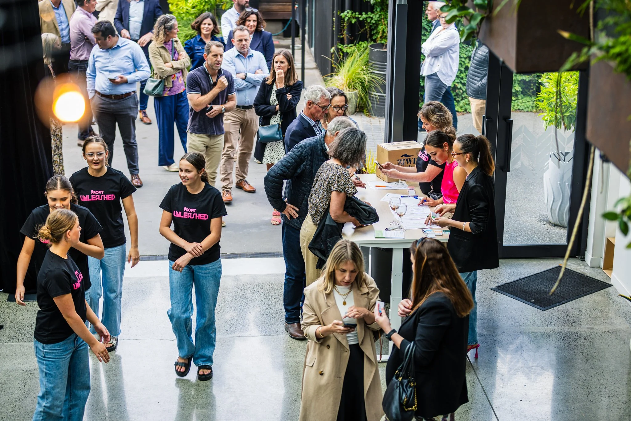 A group of people gather inside and outside a building for an event. Several women are at a registration desk, while others are standing in line or talking. Four young girls wearing black T-shirts with pink lettering are smiling and chatting in the f