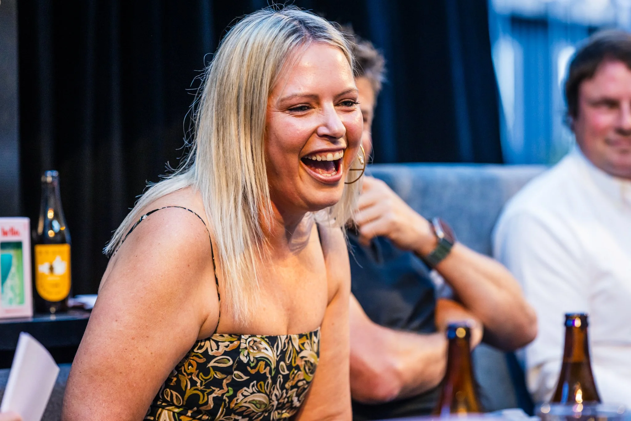 A woman with blonde hair laughing and enjoying a moment at a social gathering, seated next to two men, with bottles on the table.