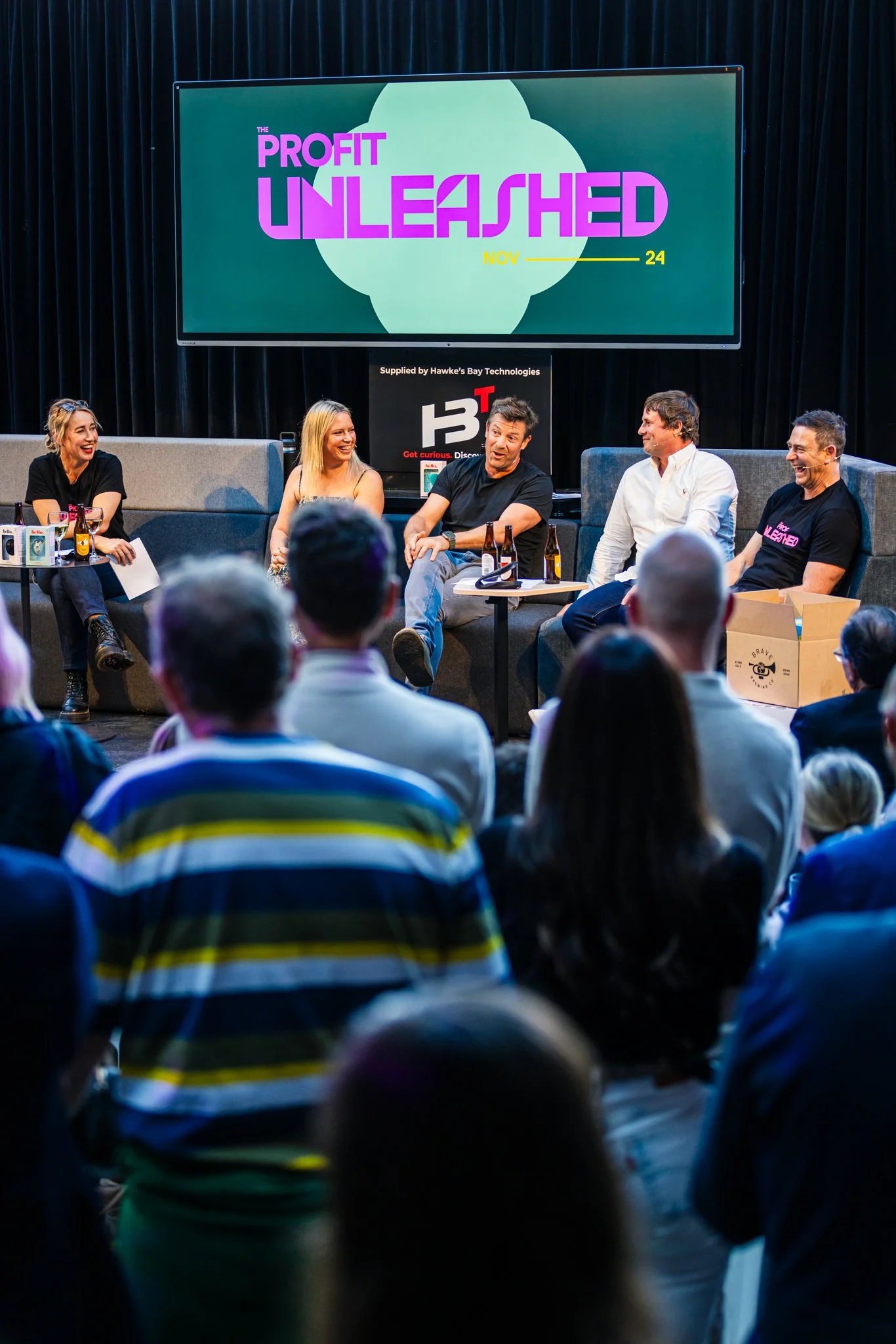 A panel discussion on stage with five people seated, and an audience in front, under a large screen displaying 'The Profit Unleashed'. The panel is engaged in conversation, with wine bottles and snacks on the tables.