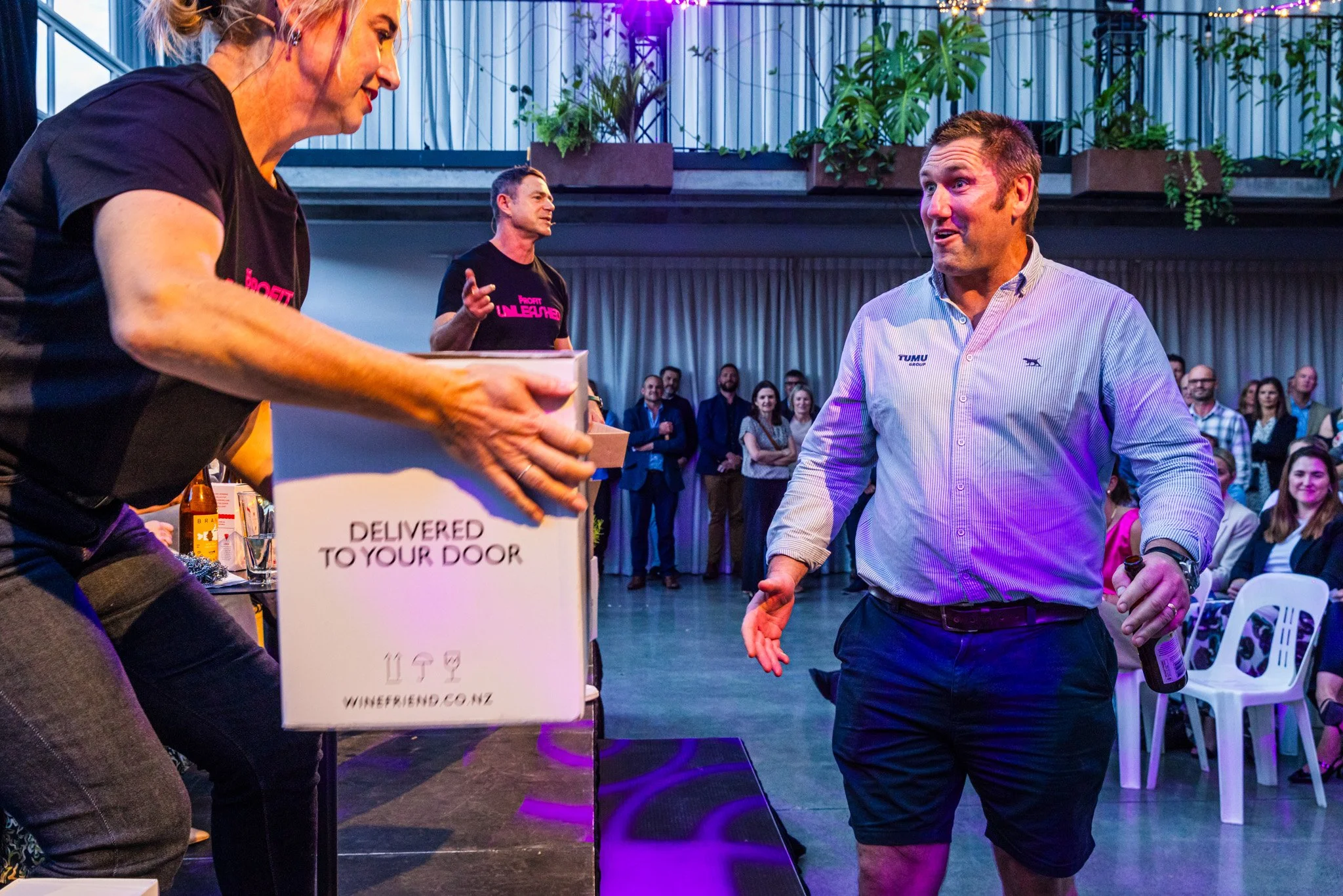 A man in a light blue shirt and black shorts receiving a box with the text 'Delivered to your door' at a corporate event, with a woman handing him the box and a person in the background speaking into a microphone. Several attendees in the background 