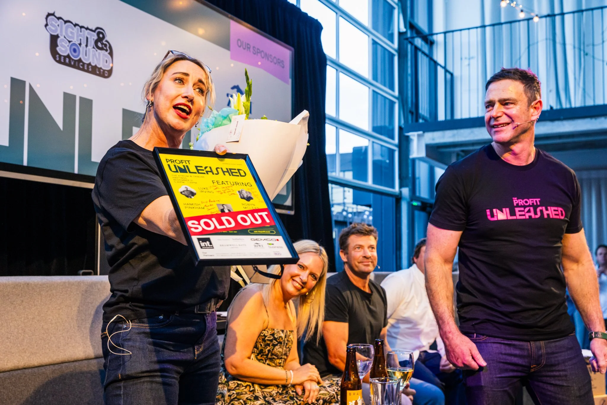A woman in black holding a bouquet and a sign that reads "Profit Unleashed Sold Out" is giving a speech or presentation at an event, with several seated people on stage and a large screen displaying event branding and sponsors behind her.