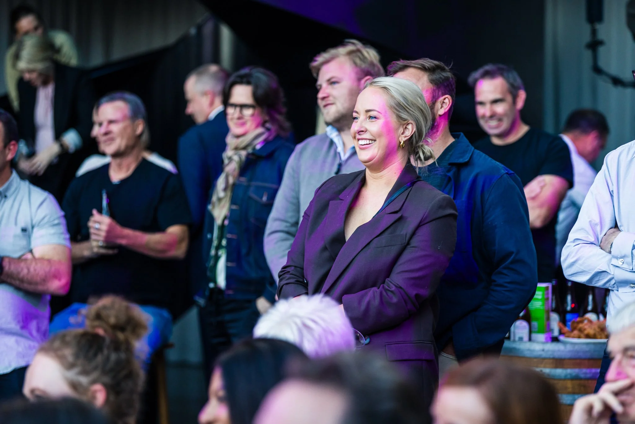 A woman in a black blazer smiling and standing among a group of people at a social event or gathering.