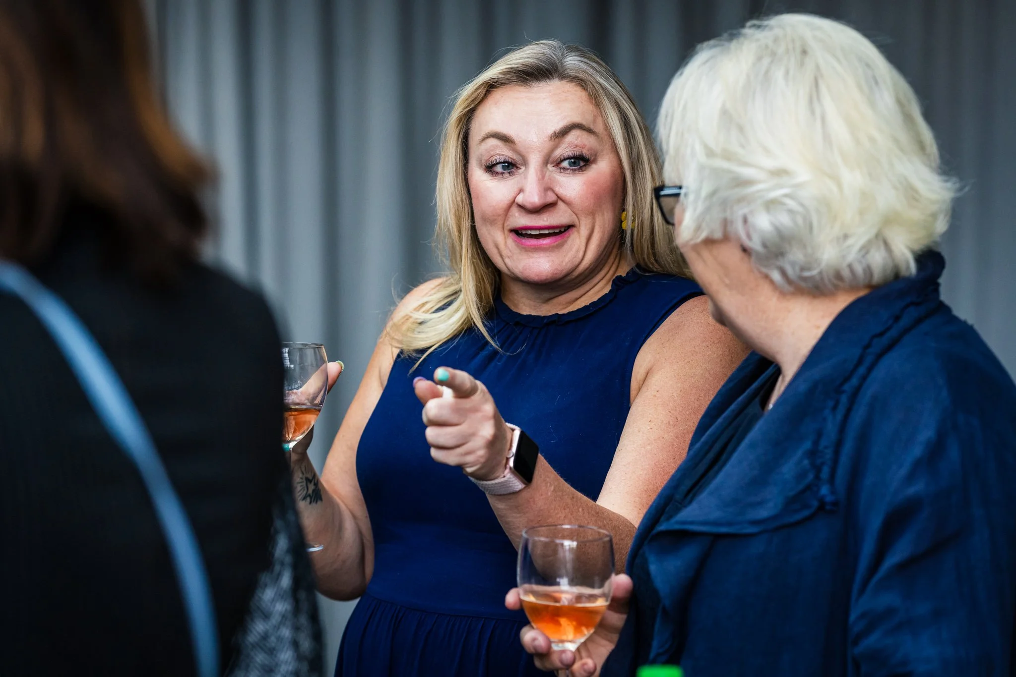 A woman in a blue dress is speaking to two other women at a social gathering, holding a glass of rosé wine.