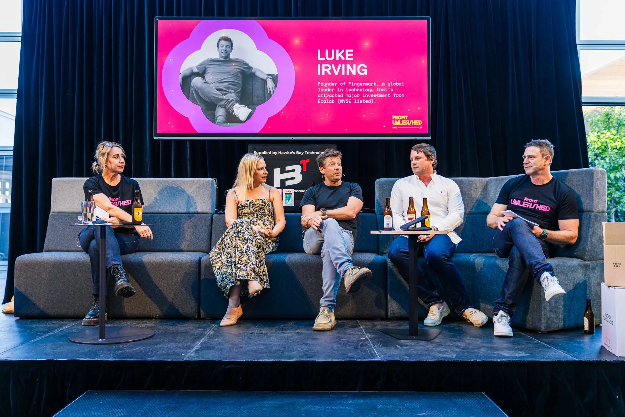 Panel of five people seated on a stage during a discussion, with a large screen behind them displaying an image of Luke Irving and event information. There are drinks and bottles on small tables next to the panelists.