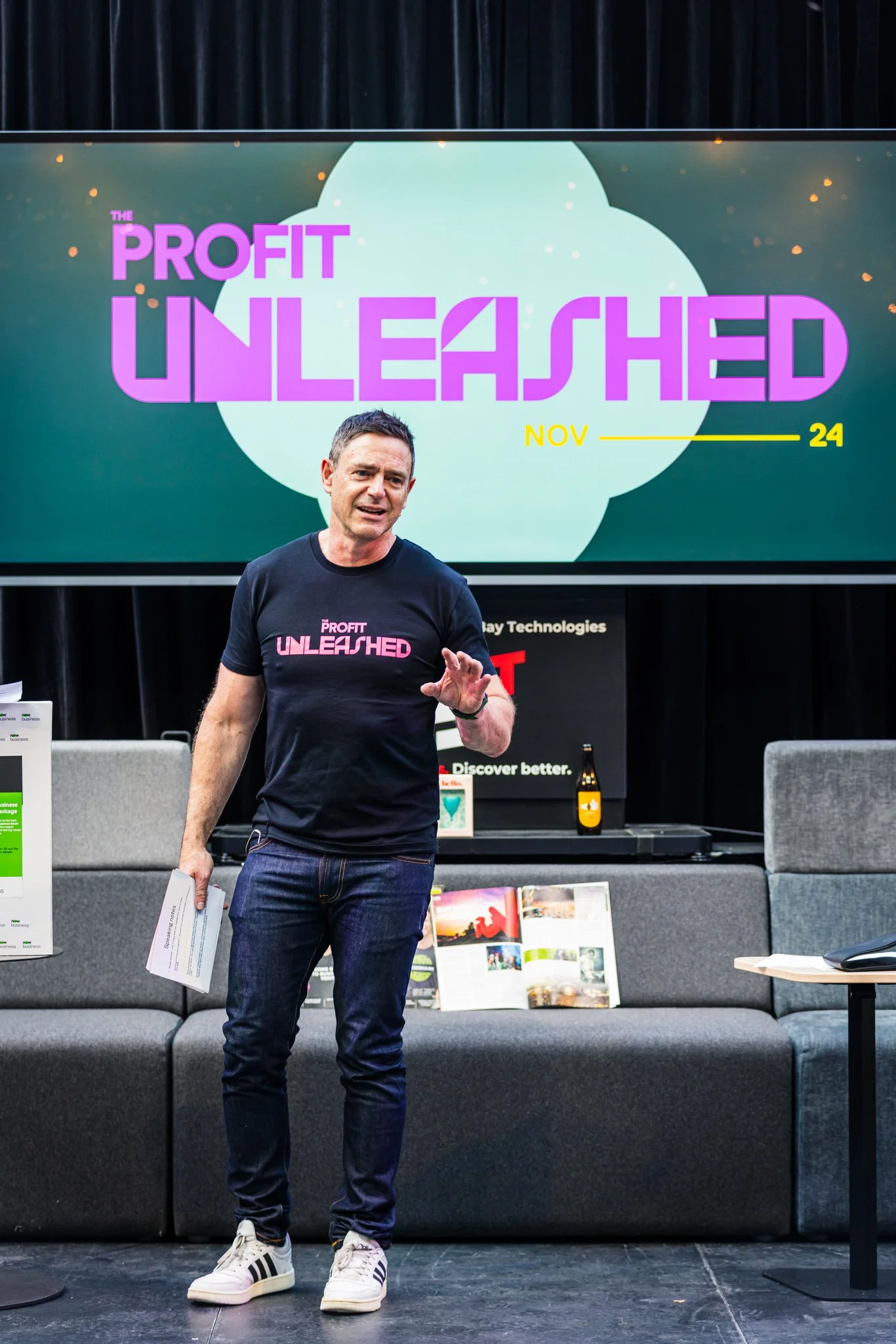 A man in a black t-shirt and jeans stands in front of a large screen displaying the words 'The Profit Unleashed' in purple and pink text, with the date 'Nov 24' in yellow. He is holding a white paper and appears to be speaking or presenting.