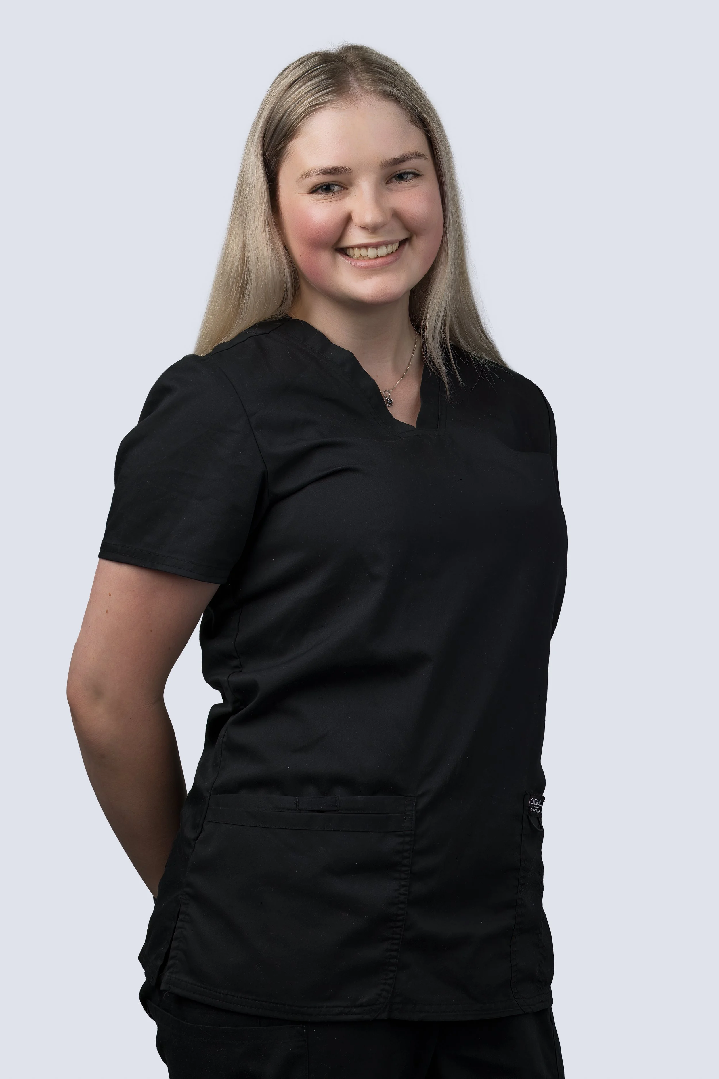 A young woman in black medical scrubs smiling at the camera with her arms behind her back.
