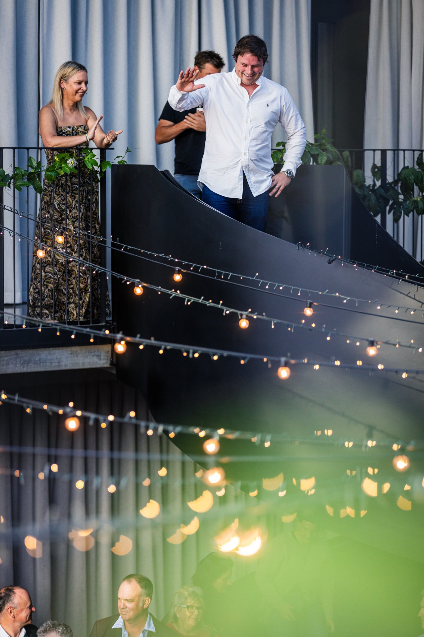 People at an indoor event, one man smiling and waving, standing on a balcony with string lights, other guests below engaging in conversation.