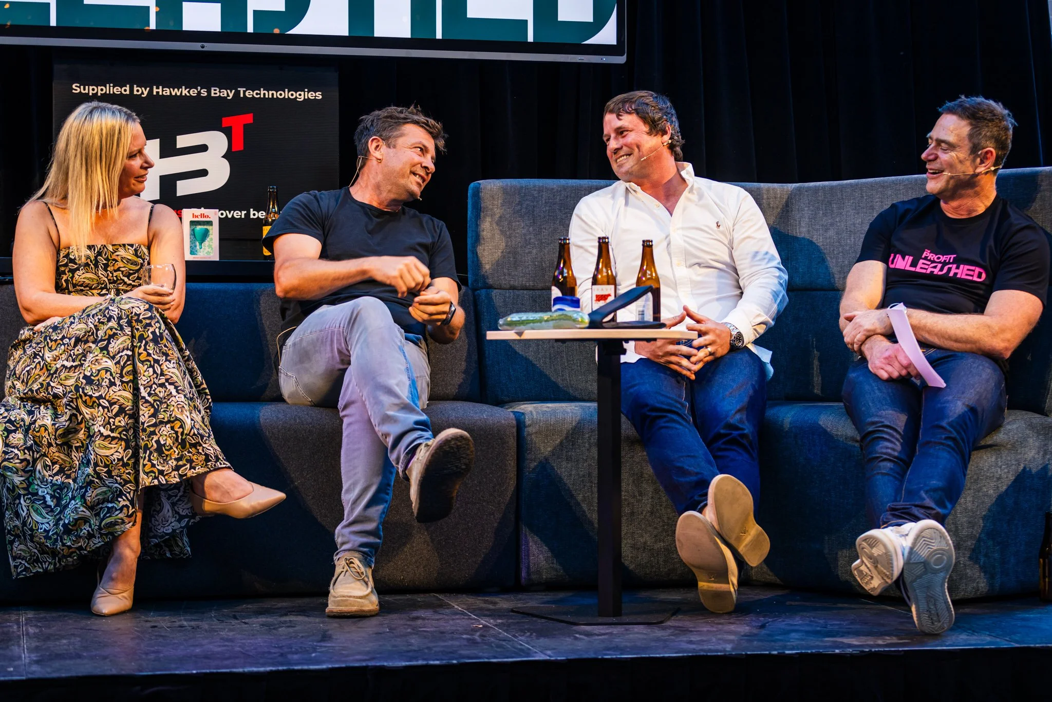Four people sitting on a dark gray sofa on a stage, engaging in conversation and smiling, with drinks and a small table in front of them. The background includes a large screen displaying logos and text.