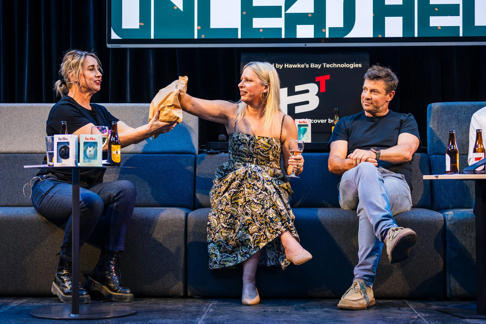 Three people sit on a stage with a black curtain background. The woman in the middle is exchanging a paper bag with the woman on the left, who is handing it to her. The woman in the middle is holding a glass of wine, while the man on the right observ