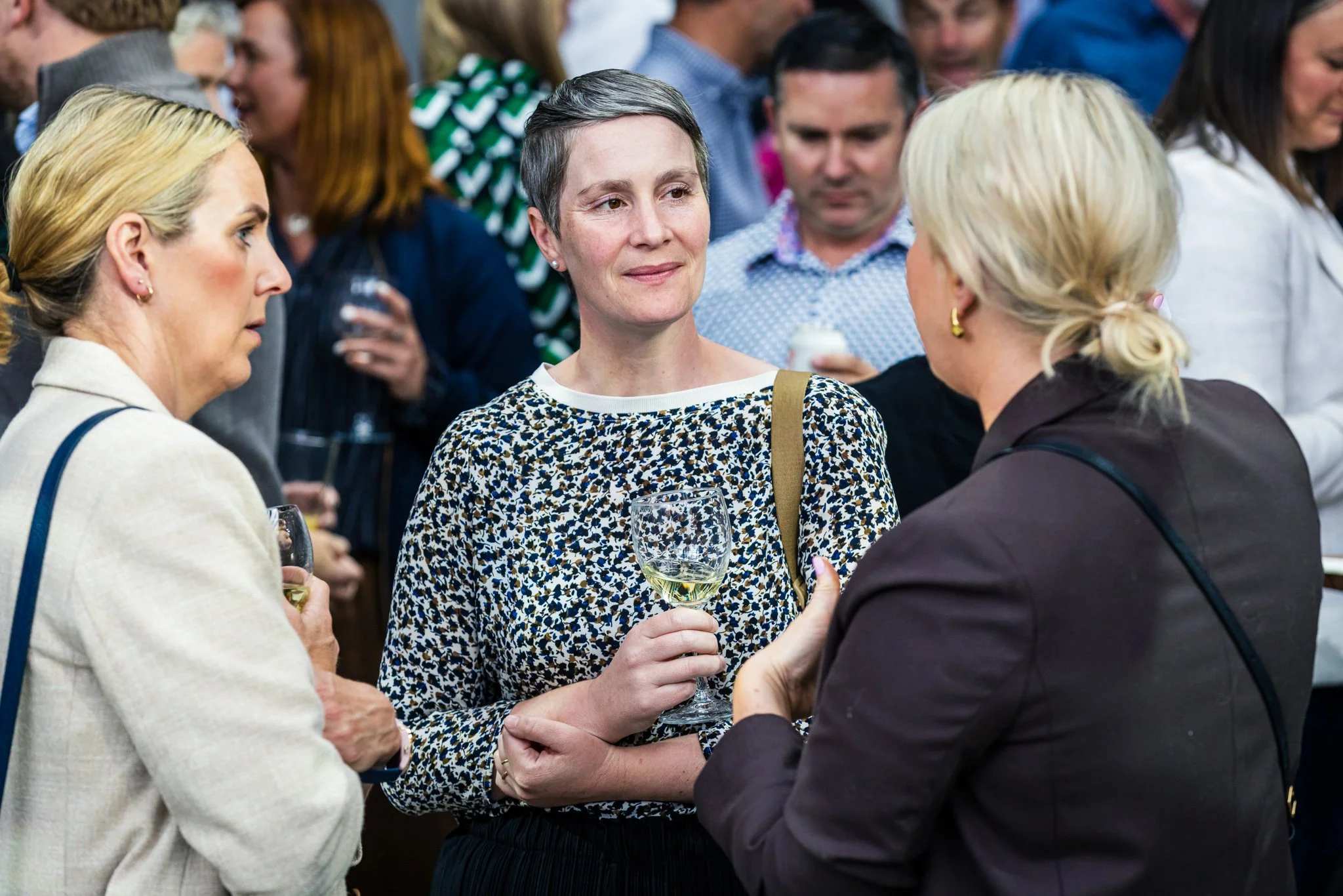 Three women having a conversation at a social event, with others mingling in the background. One woman is holding a wine glass, and they appear engaged in discussion.