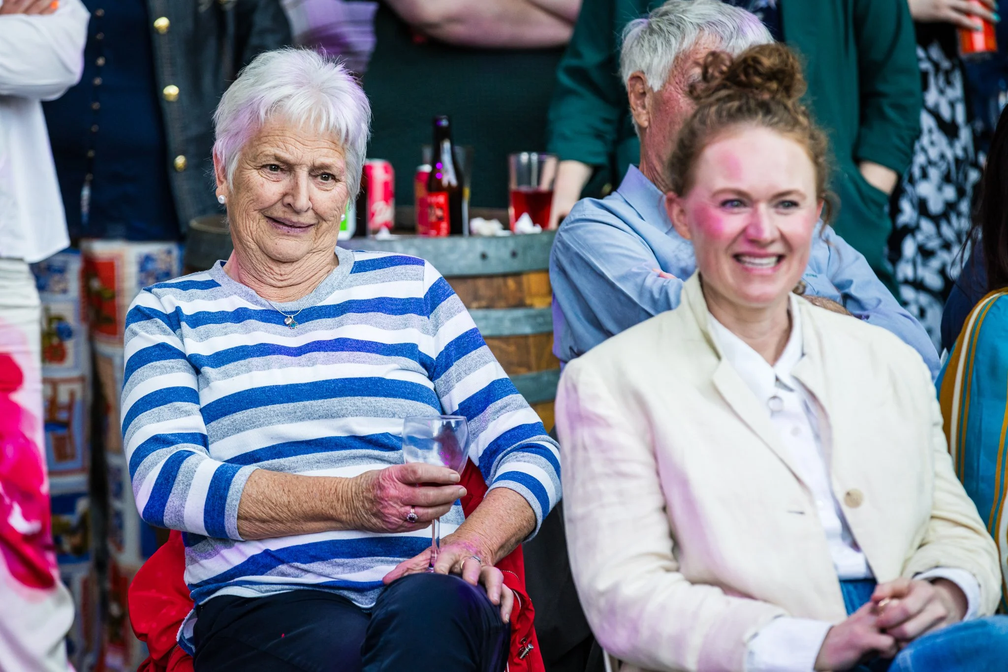 People at a social gathering, with elderly women and a mousy woman smiling, holding a wine glass, in an outdoor setting with drinks and decorations.