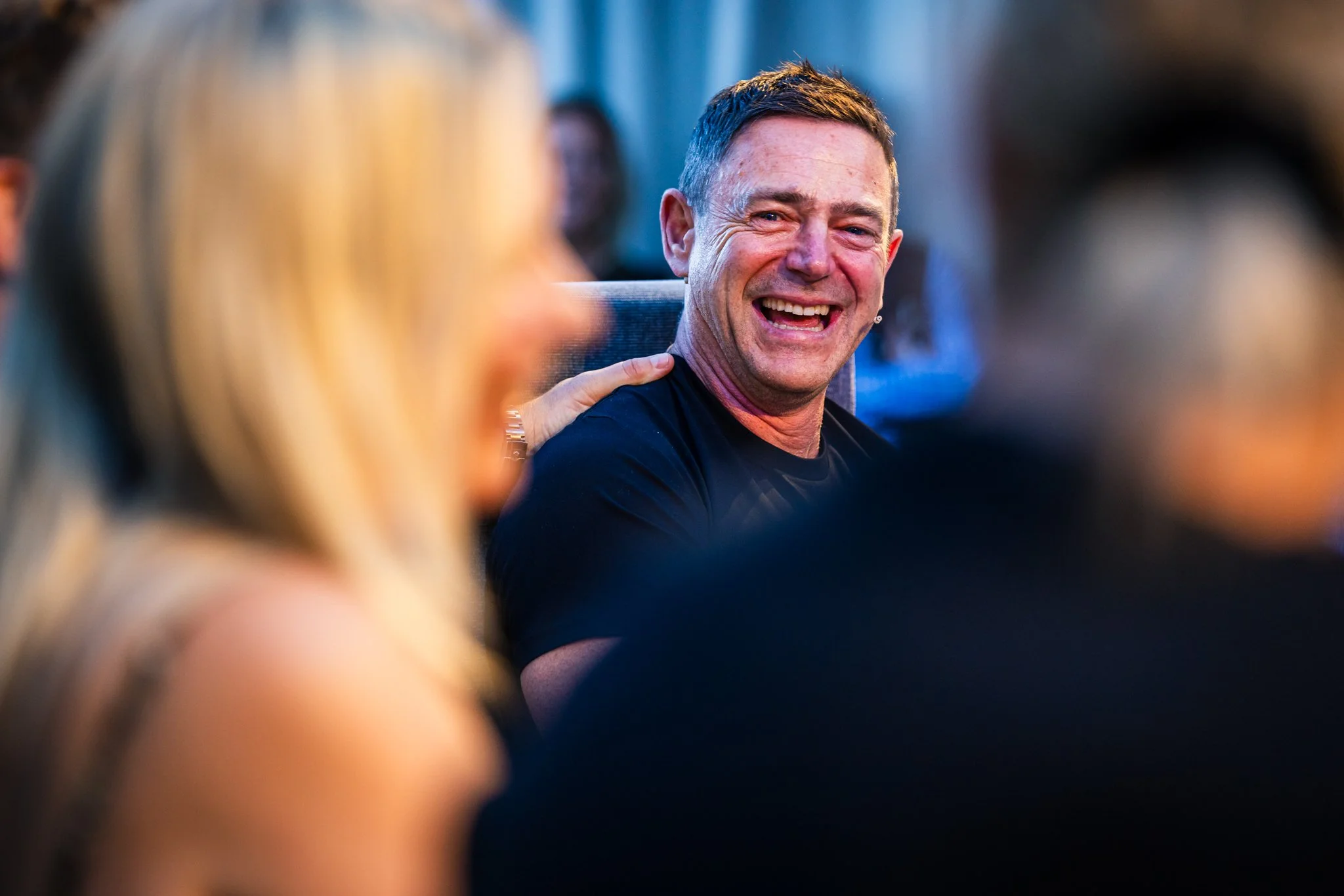 A man with short dark hair smiling and laughing at a social gathering, surrounded by blurred people.