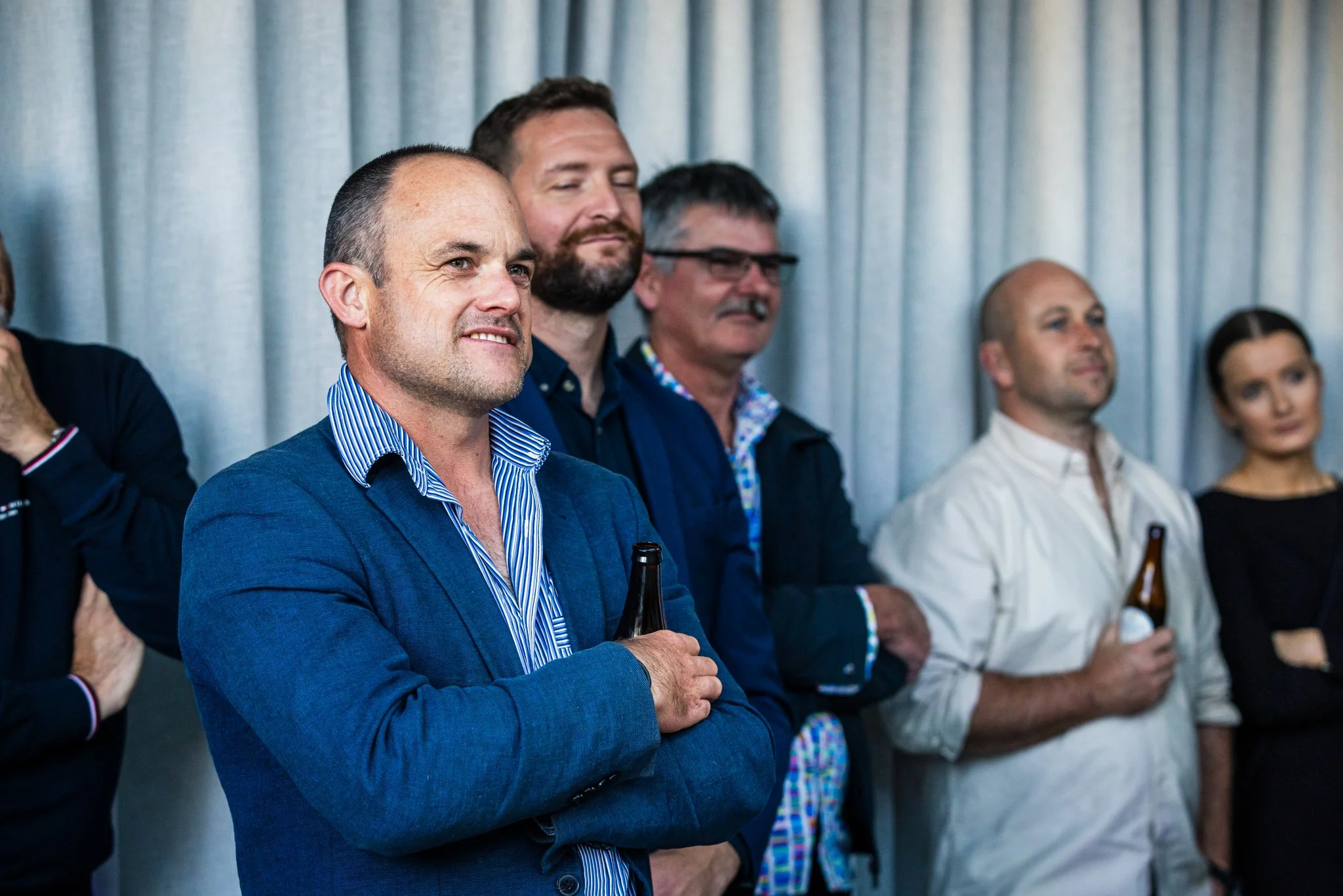 Group of people at a social event, standing in front of a curtain, some holding bottles of beer, engaging in conversation.