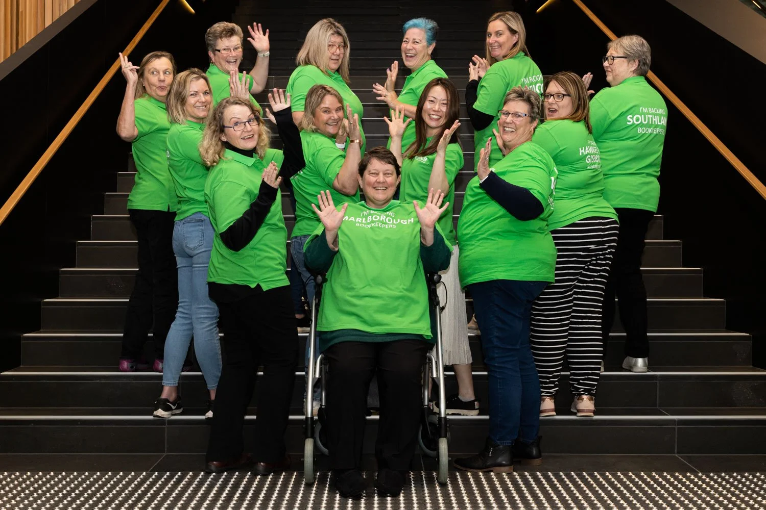 Group of women wearing matching green t-shirts, posing on stairs, some smiling and waving, one woman in a wheelchair with hands raised, all looking happy and excited.