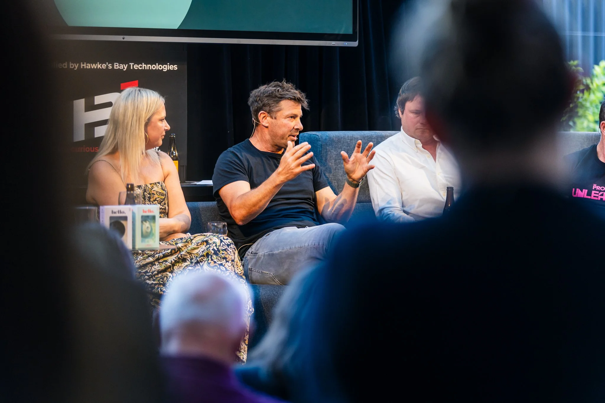A panel discussion with four people, including a man in a black T-shirt speaking animatedly, a woman in a patterned dress, and two men in white shirts, sitting on a stage with a black curtain background and a screen overhead.