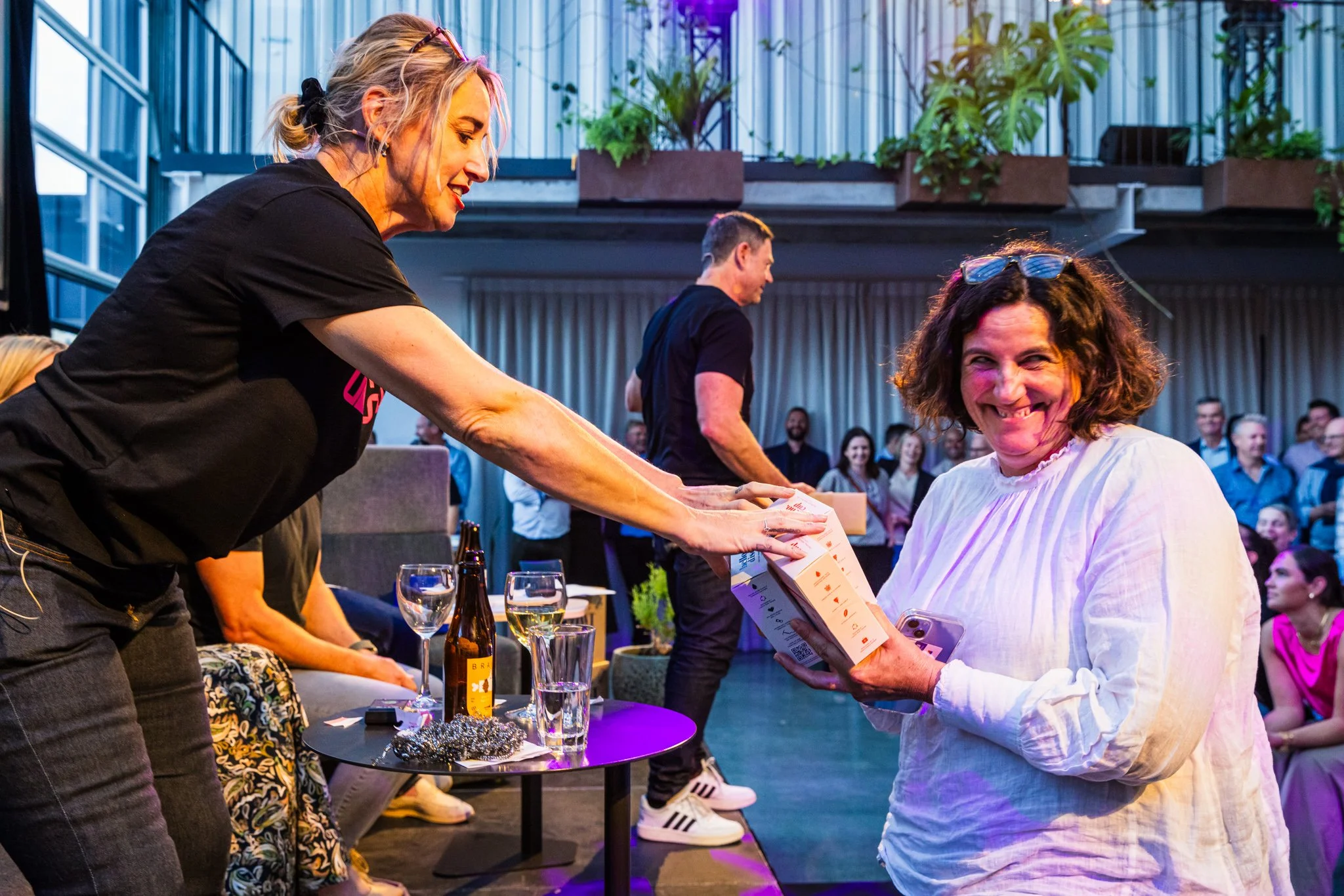 A woman in a black T-shirt is handing a boxed gift to a smiling woman in a white blouse at a social event, with a crowd of people in the background.