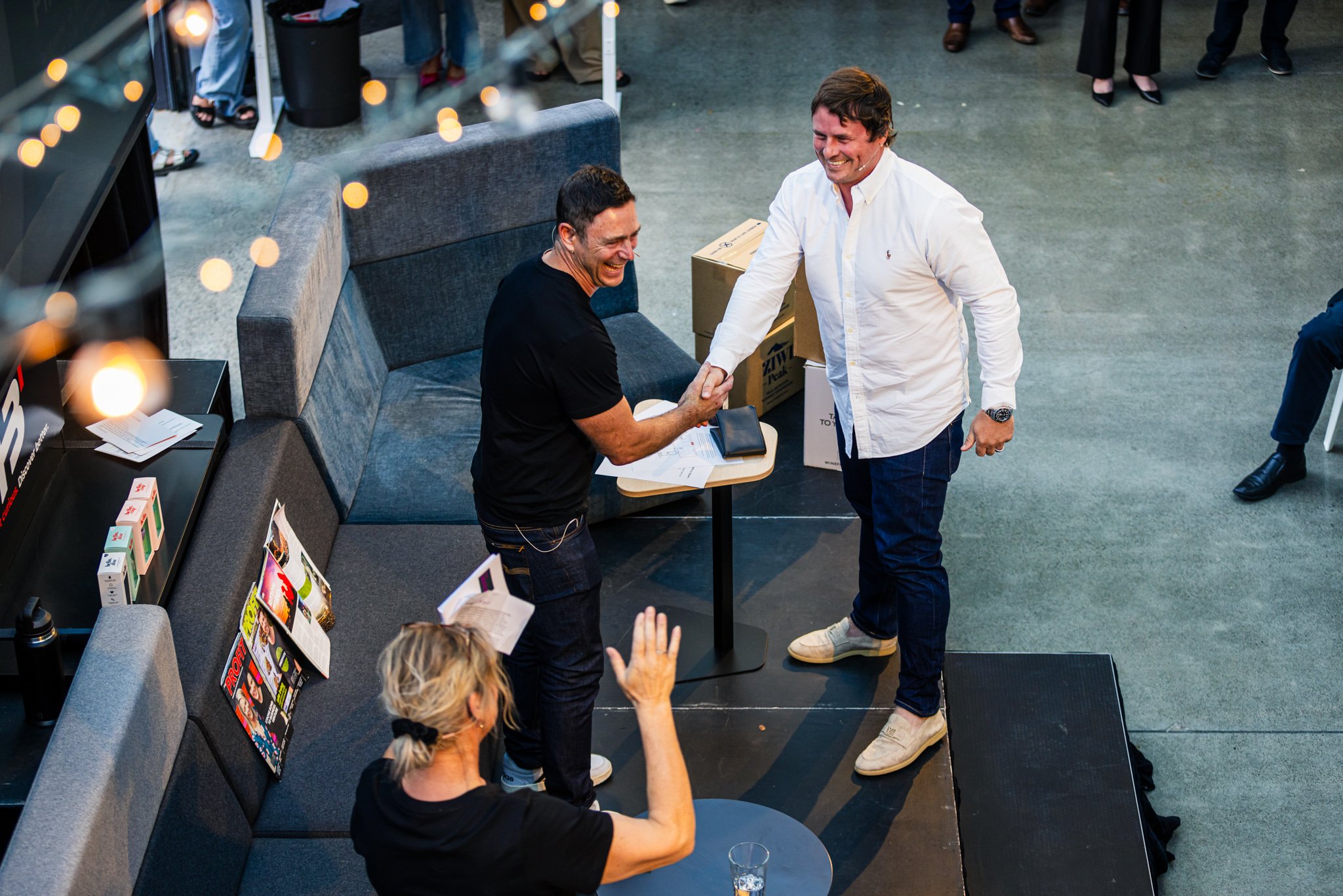 Two men shaking hands and smiling at each other at a social event, while a woman seated at a table in front of them raises her hand in greeting or farewell.