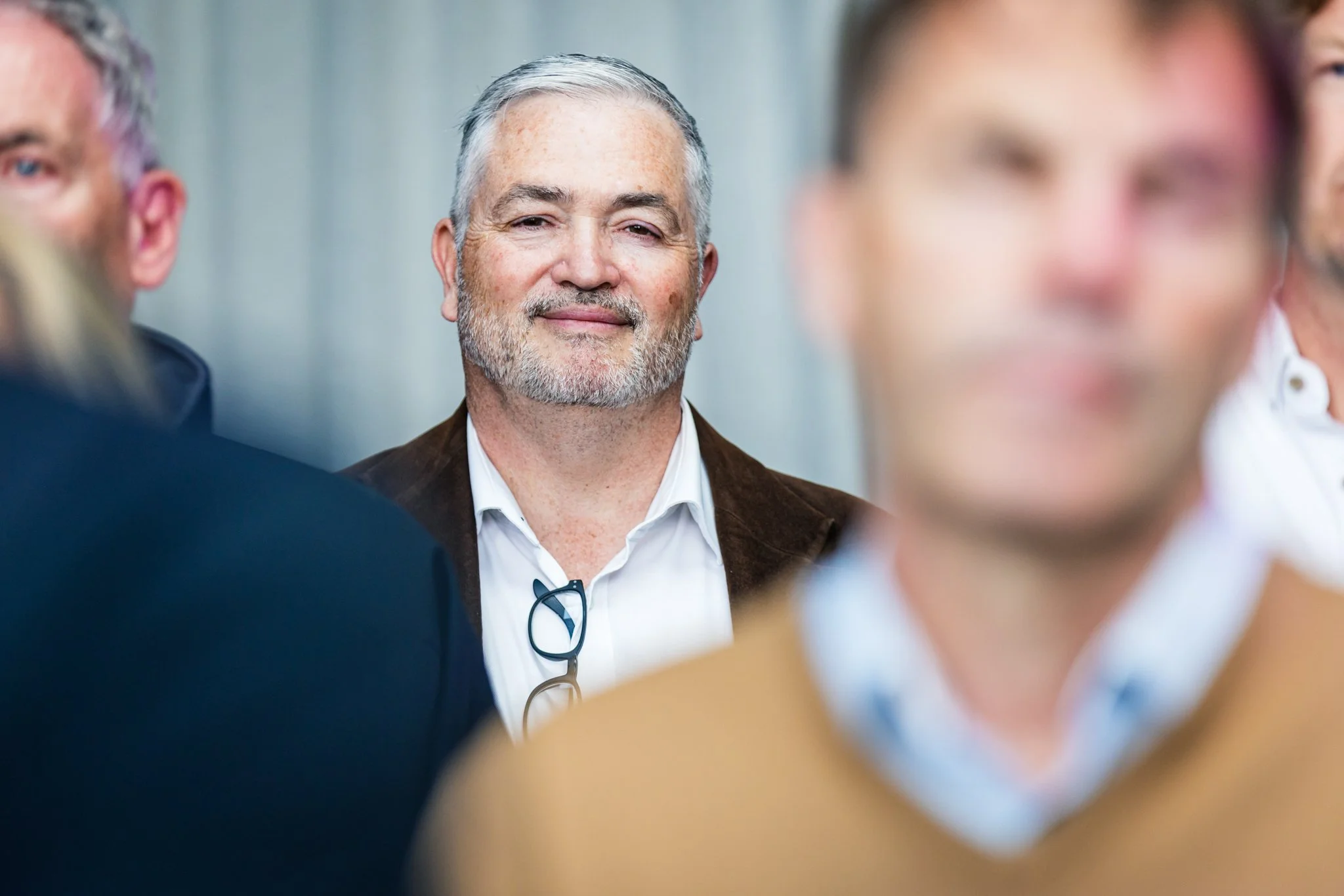 Smiling older man with glasses hanging on his shirt, surrounded by people in a professional setting.