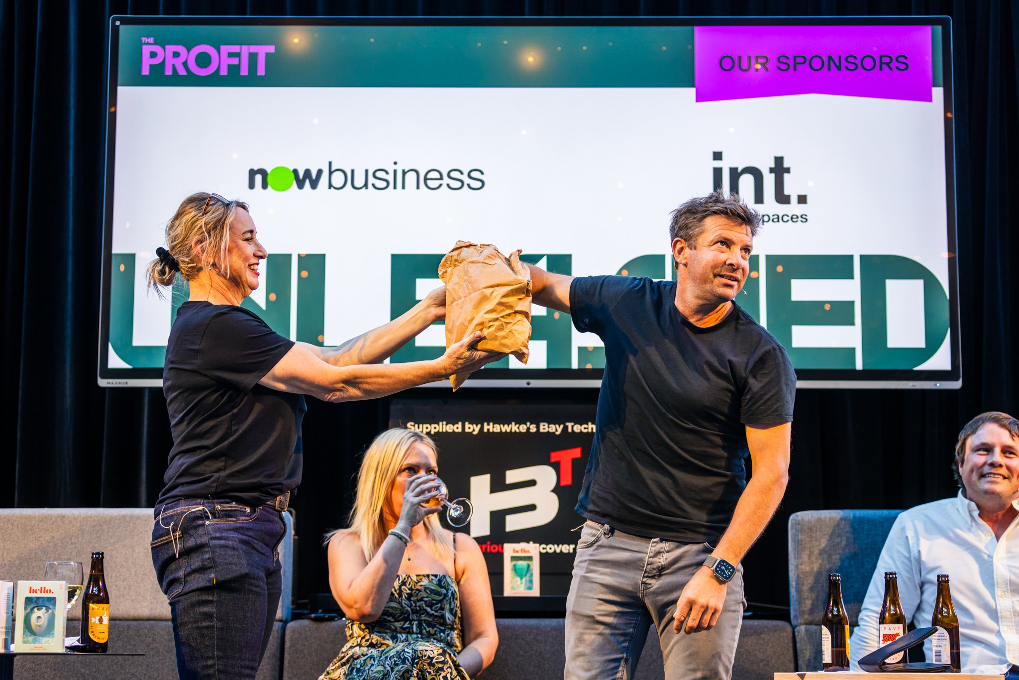 A man receiving a paper bag gift from a woman on stage during a panel event, with two seated women and drinks on the table, large screen behind displaying sponsors and event branding.
